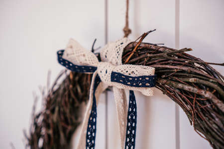 Woven brown hanging natural tree wreath from wickers with decorative white bow tape lace. White door and wall as background. Traditional seasonal ethnic holiday decoration. Close up. Copy spaceの写真素材