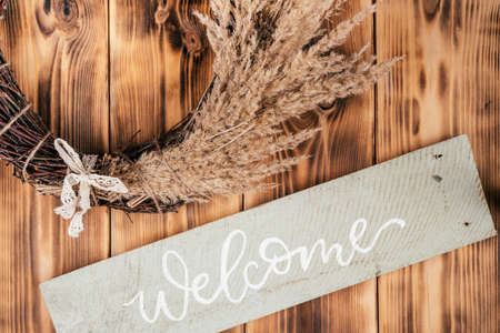 Dry herbal, cereal tightly woven wreath with string and nameplate welcome. Rustic brown wooden and planking door on background. Celebration and preparation, warm greeting of guests. Cosy compositionの写真素材