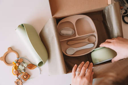 Cropped woman hands packing and wrapping shipment with silicone baby dish, plate, spoons, bowl, bib,...