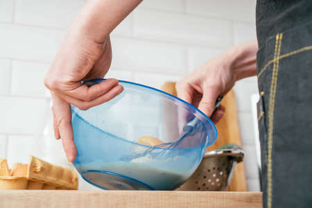 Bottom view of unrecognizable man in black kitchen apron beating eggs in blue plastic bowl with wire...