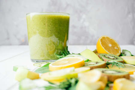 Partially blurred glass of green smoothie with green and yellow fruits and herbs cut into pieces on white table. Ingredients for smoothie. Slices of lemon, apple, kiwi, cucumber, spinach and tarragonの写真素材