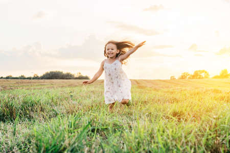 Portrait of smiling girl playing, jumping and running on grass hay field paths of dry grass in the sunset. Waving hands. Forest on bright light background. Cloudy sunny sky. Haying timeの写真素材