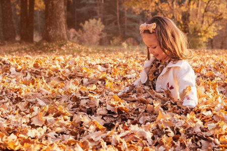 Little girl 3-4 years old sits outdoor in park in rays of setting sun with feet buried under oak...