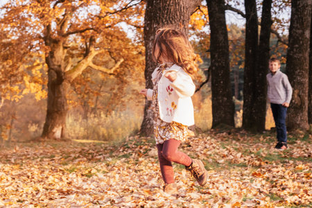 Little girl 3-4 years old runs from boy brother 9-10 years old on autumn leaves outdoor near large thick oak trunks in park in rays of setting sun. Siblings play catch up outdoorsの写真素材