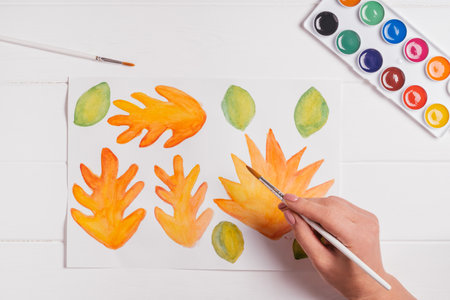 Female hand drawing autumn leaves of different shapes, sizes and colors with watercolors and brush on white wooden table. Top view. DIY handmade autumn decorationsの写真素材