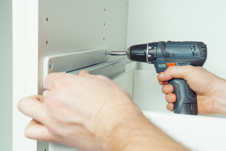 Male hands hold cordless electric screwdriver, screw to fix rail, assembling drawer of white dresser or wardrobe. Self-assembly of furniture at home, DIY, furniture repair and rearrangementの写真素材