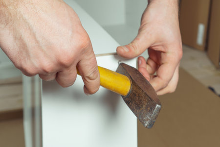 Male hands hold metal hammer with yellow plastic handle, driving dowel, assembling white drawer of dresser or wardrobe. Self-assembly of furniture at home, DIY, furniture repair and rearrangementの写真素材