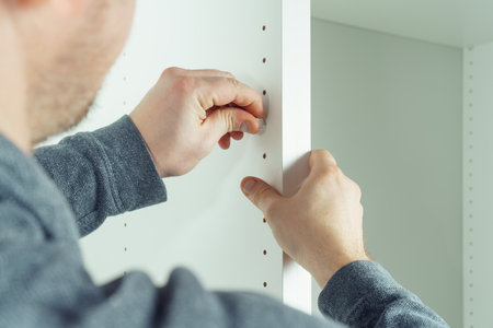 Male hands insert plastic support into one of series of holes to fix white shelf, assembling dresser or wardrobe. Self-assembly of furniture at home, DIY, furniture repairの写真素材