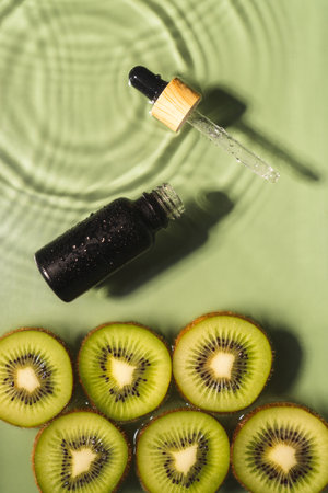 Facial pipette serum black bottle, whey beauty product near fruit tropical kiwi slices in water splashing fresh transparent, flecks. top view. Organic skin face and body moisturizing. Verticalの写真素材