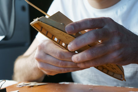 Hands of man gluing plywood details for ship model with glue, holding with fingers. Process of building toy ship, hobby, handicraft. Table with various materials, parts and devices for workの写真素材