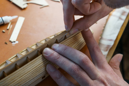 Hands of man gluing plywood details for ship model with glue, holding with fingers. Process of building toy ship, hobby, handicraft. Table with various materials, parts and devices for workの写真素材