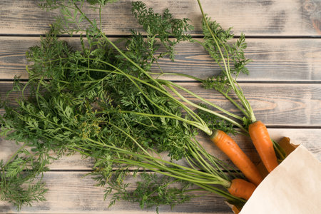 Bunch of fresh ripe carrots with long green haulm on faded wooden table, top view. Unpacking vegetables from eco paper bag. Organic food or healthy diet concept. Objects layout. Planking.の写真素材