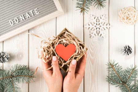 Female hands hold present donation box with red paper heart postcard, top view. Decorative spruce...