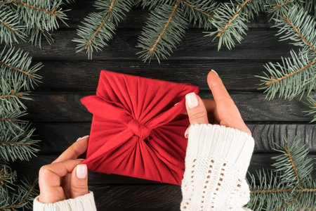 Female hands in white sweater tie knot on christmas present in furoshiki package, top view. organic spruce branches frame. Ecological Christmas or New Year holiday concept, eco decor, zero waste.の写真素材