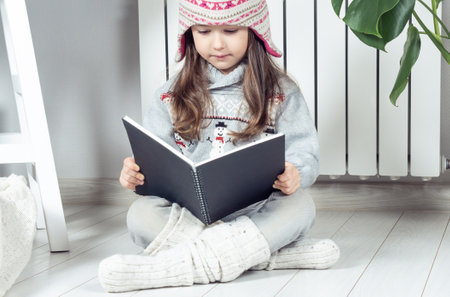 Pensive, serious, cute little girl in woolen hat and warm clothes with snowman reading book or...
