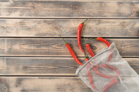 Red hot tasty chili peppers in ecological degradable string bag on wooden background. Spicy food, seasoning and condiment. Recycling and sustainability development, lifestyle. Top view, copy spaceの写真素材