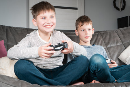 Portrait of two smiling glad teenage boys sitting on grey sofa at home, holding gaming controller...