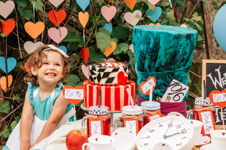 Little smiling girl 2 years old dressed as Alice in Wonderland near table with fairy tale props. Cake, Hatters hat, inscriptions Eat me and Drink me, clock, card suits. Themed childrens book birthdayの写真素材