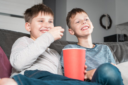 Portrait of two laughing positive teenage boys sitting on sofa at home, watching funny movie comedy video cartoon on TV, eating popcorn from red bucket having fun. Free time, friendship, relationship.の写真素材