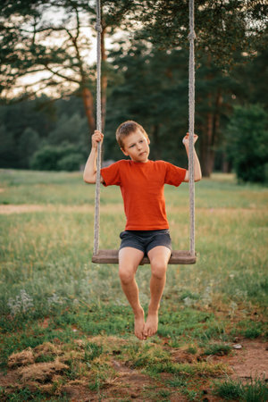 Barefoot boy 7-10 in red t-shirt with bruised legs sitting on rope swing in countryside among pines, in rays of setting sun. Verticalの写真素材