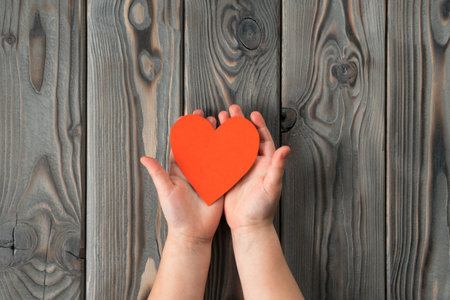 Unrecognizable open hands holding and showing small red paper heart on dark wooden background. I give you my heart and soul. Family and children day or valentines day celebration. Copy space, top viewの写真素材