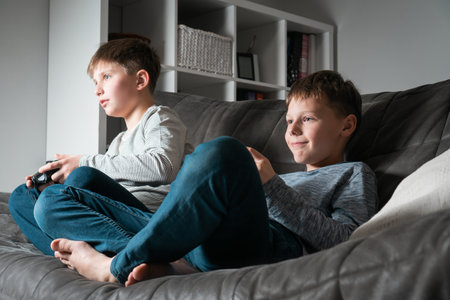 Cute school aged boys sit on sofa with game joysticks, from below view. Children play videogames. Happy kids relaxing in home room at daytime together. Modern electronic games. Best friends lifestyle.の写真素材