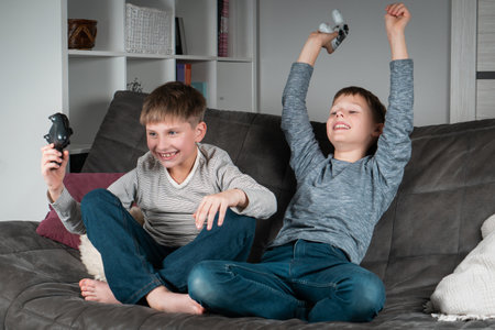 Overjoyed teenagers sit on sofa with game joysticks. Cheerful boys play videogames. Kids having fun...
