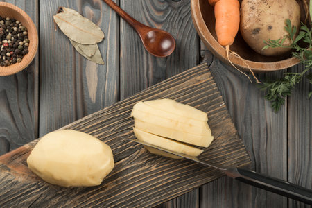 Cutting into slices raw yellow potato with knife on kitchen wooden board. Fresh sliced potatoes, traditional cuisine. flat lay view. Ripe vegetables and flavorings. Prepare ingredients for cooking.の写真素材