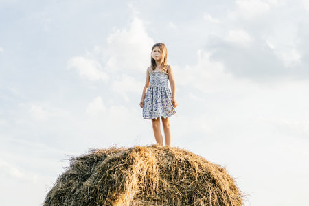 Pretty little girl stand at the top of haystack looking away. Full length photo, from below view. Resting on hayrick. outdoor walking. beautiful blue sky. freedom. Summer vacation, countryside lifestyle.の写真素材