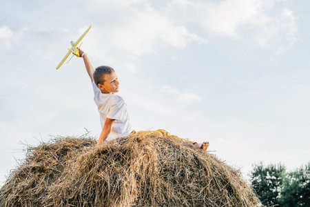 Happy school aged boy sitting at top of haystack launch toy airplane. Carefree child on haymaking play toys. outdoor walking. beautiful blue sky. Summer vacation, countryside lifestyle, haying.の写真素材