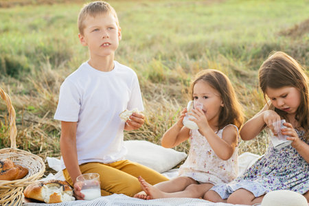 Happy friends eat fresh buns and drink milk sitting on blanket on field. Portrait of little children resting on grass. Tasty and nutritious outdoor picnic. Summer vacation, countryside lifestyle.の写真素材