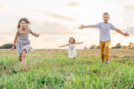 Happy little children joyfully run forward across field waving arms wide. Carefree kids on grassy meadow. outdoor walking. beautiful sunny sky. Summer vacation, countryside lifestyle.の写真素材