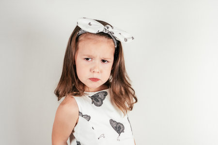Unhappy little girl with offended facial expression. Portrait of discontent child on white. Kid in beautiful festive dress and headband. Saint Valentine's Day concept, holiday of all lovers.の写真素材