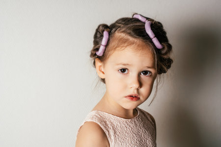 Adorable upset little girl with hair curlers. Portrait of sad child on white. Kid in beautiful festive dress prepare hairstyle. Saint Valentine's Day concept, holiday outfit, beauty care.の写真素材