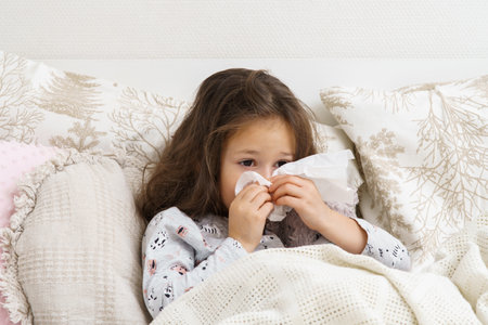 Little girl blow stuffy nose with white handkerchief. Portrait of unhealthy child. Kid in sleepwear lie under blanket and have bed rest. Home treatment. Runny nose, snot, cold symptoms, cure.の写真素材