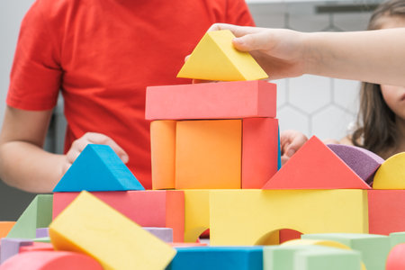 Cropped photo of preteen children friends sitting at table in kitchen, building tower house castle of colorful wooden construction blocks geometric figures. Board games, entertainment, development.の写真素材