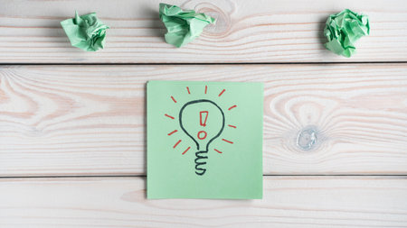 Flat lay of green sticker note with light bulb drawn with black marker and red exclamation mark inside near crumpled notes on grey wooden table. Idea, creative work, brainstorming, invention, genius.の写真素材