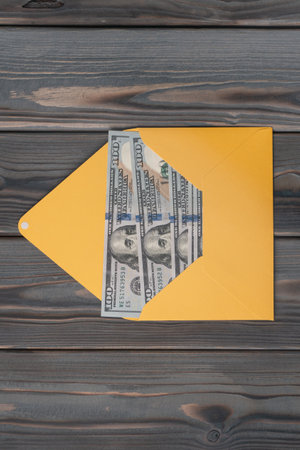 Flat lay of open yellow envelope with hundred dollars banknotes bills on grey wooden table....