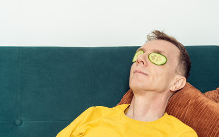 Happy man lying on sofa with eye patches from fresh cucumber slices in living room. Adult person reclining on pillow with mask for dark circles under eyes. Home relaxation, beauty treatment.の写真素材