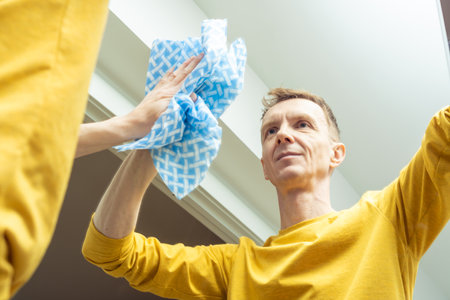 Adult man wiping room mirror with dry rag, from below view. Happy man rub from dust and polish mirror with household cloth. Domestic life, tidy housekeeper. Housework, home cleaning concept.の写真素材
