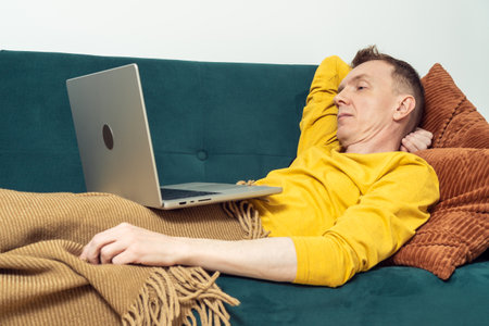Tired man lie on sofa and watch movie on laptop in living room. Adult person reclining on pillow...
