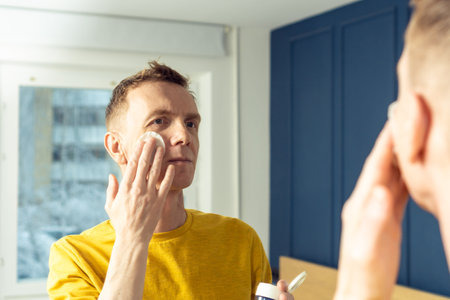Mature man apply face tonic with cotton pad, then massage with hands. Male portrait in room mirror. Skincare morning routine, careful beauty treatment, home lifestyle. Face cleansing, washing.の写真素材