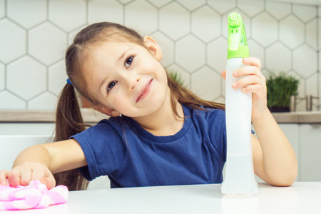 Happy little girl with detergent sprayer and household rag at table. Portrait of child tidying up kitchen, home cleaning concept. Kid help clean up house. Room cleaning supplies, cleanser.の写真素材