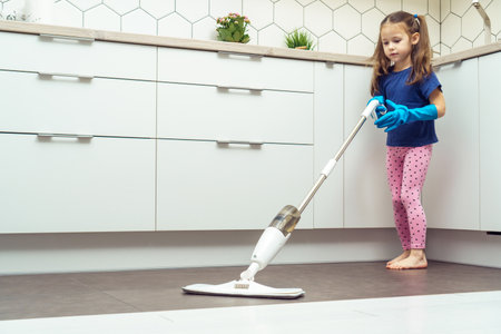 Pretty little girl in household gloves wash floor by modern mop with removable brush and water container near kitchen set. Full length photo of child tidying up kitchen side view. Home wet cleaning.の写真素材
