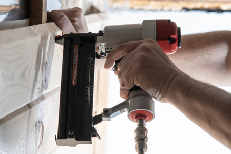 Cropped male builder hands use pneumatic stapler gun for clipping wooden timber board. Scoring metal nails and staples, making layout on wooden plank. Builder and civil engineer occupation. Side viewの写真素材