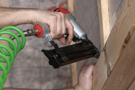Unrecognizable cropped male hands using pneumatic nailer, nail gun with spiral cable for wooden timber board. Make layout on wooden plank. Clipping and scoring metal nails and staples. Low angle viewの写真素材