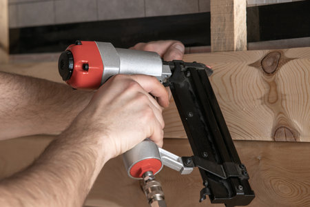 Close up cropped male hands using pneumatic nailer, stapler nail gun for wooden planks and board. Clipping and scoring metal nails and staples, making layout on wooden plank. Builder first person viewの写真素材