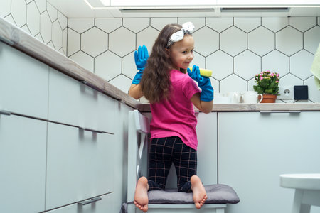 Happy little girl in protective gloves wash up dishes with sponge in kitchen sink sitting on chair. Portrait of child tidying up kitchen back view. Home cleaning concept. Stylish interior, furniture.の写真素材
