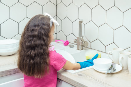 Little girl in household gloves washing up tableware with foamy dishwashing sponge and dish soap in kitchen sink under water jet. Portrait of child tidying up kitchen back view. Home cleaning concept.の写真素材