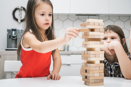 Focused, interested little long haired girl moving wooden block out of tower, playing board game with friends. Balance and stability of wooden brick tower. Educational and logical games for childrenの写真素材